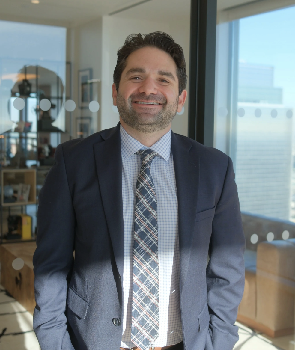 Portrait of attorney Rod N. Armande, Esq. in a navy suit and plaid tie, smiling in his downtown Los Angeles high-rise office with the city visible through the window